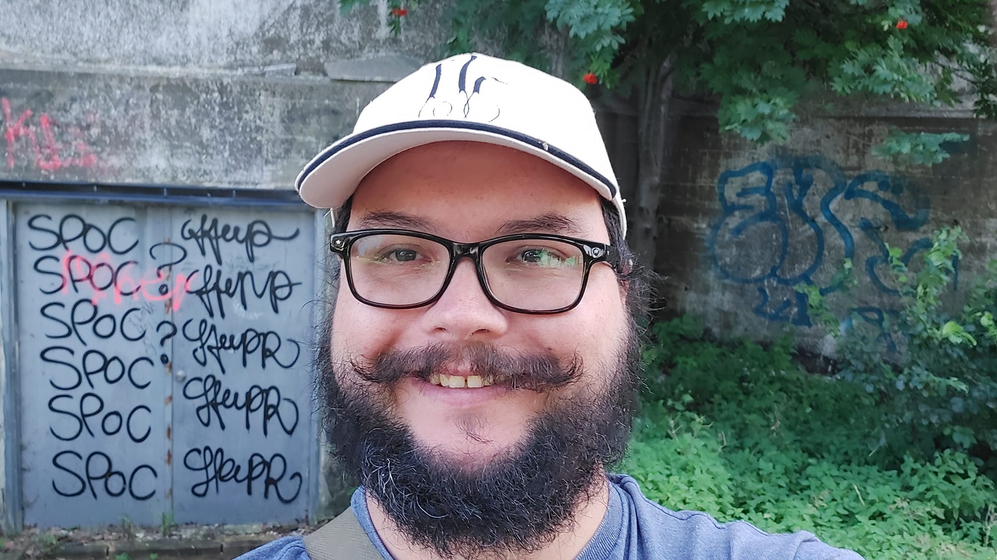 A man with short hair and bushy facial hair stands in front of a steel door covered in grafiti